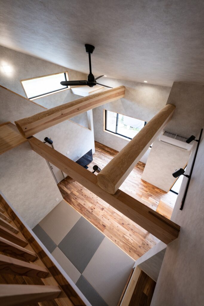 吹き抜けを2階から見下ろした室内写真|大きな梁と畳スペースがつながる新築住宅