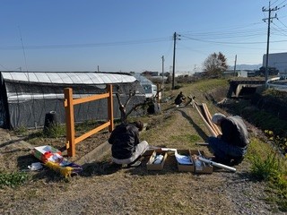 田園風景をバックに、大工3名で協力して竿に取り付ける紐などを準備している作業風景。